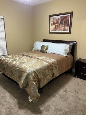A bedroom with a bed, a nightstand, a framed picture on the wall, and a window with blinds.