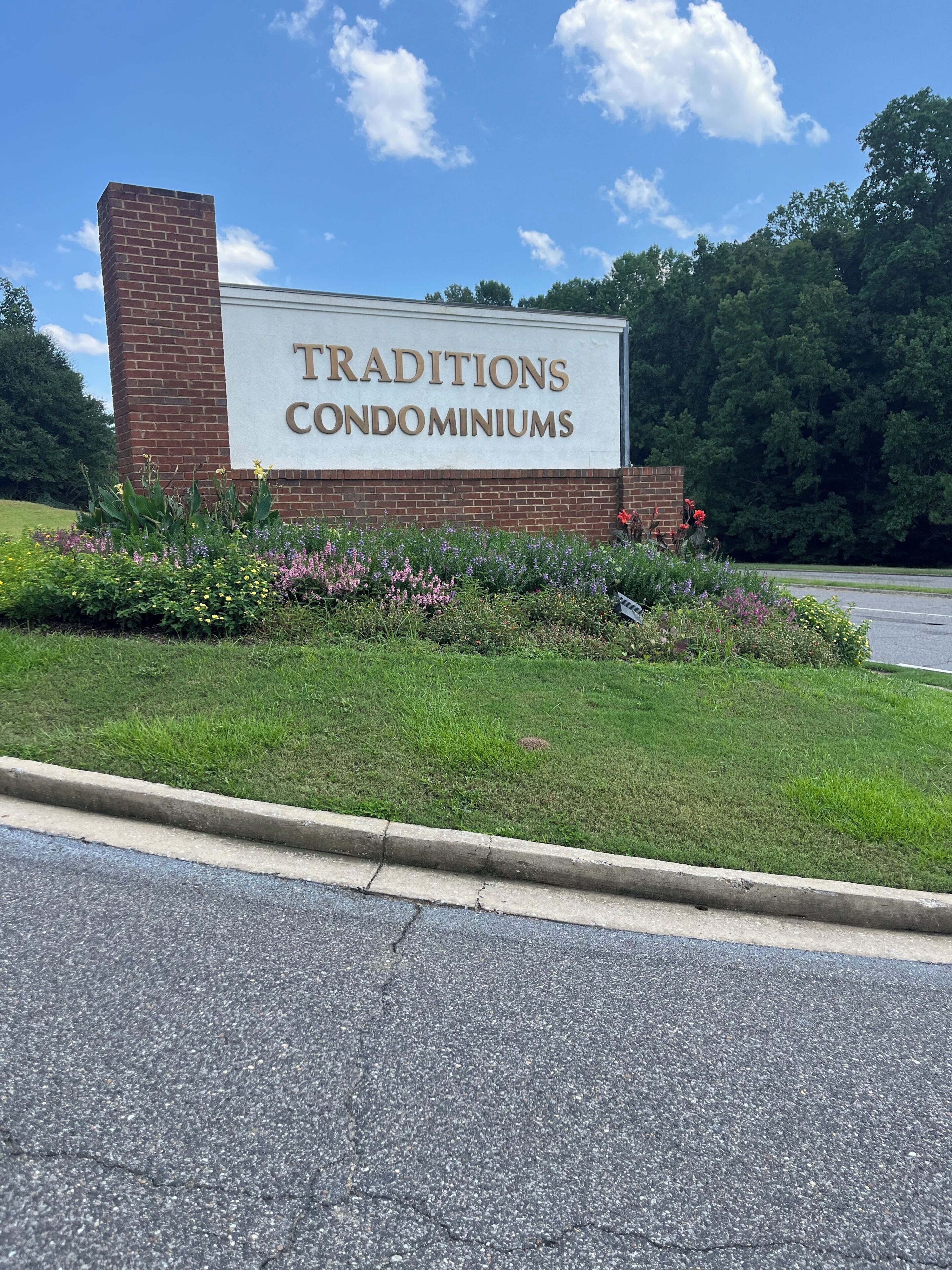 A sign for 'Traditions Condominiums' with a brick wall and a grassy area with flowers.