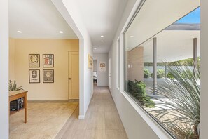 A hallway with framed artwork on the walls, a wooden table with a plaque, and a view of the outdoors through large windows. - The Beehive - A true midcentury estate (Palm Springs)