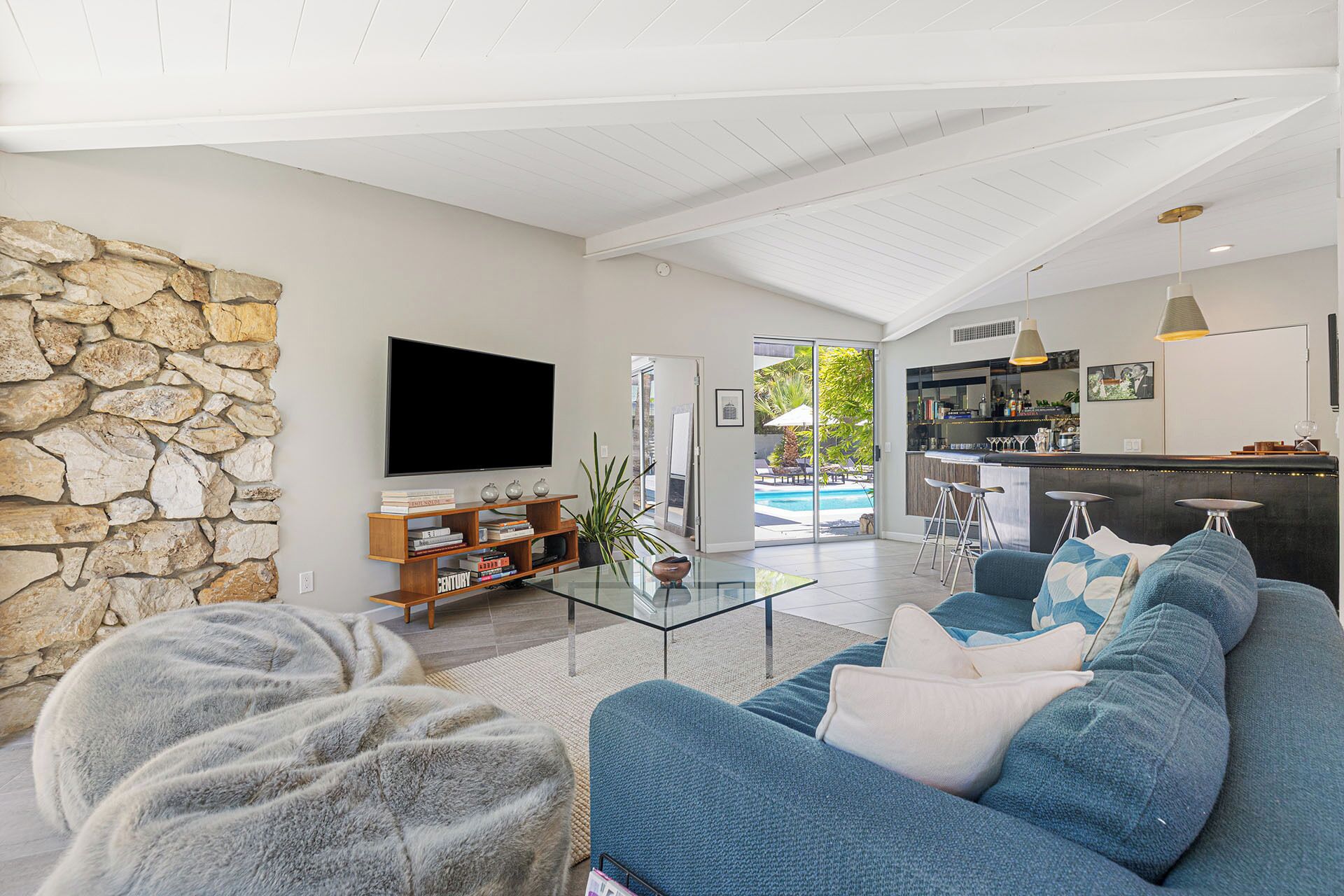 Ein geräumiger Wohnbereich mit Steinwand, Flachbildfernseher, Glastisch und blauem Sofa mit Kissen.