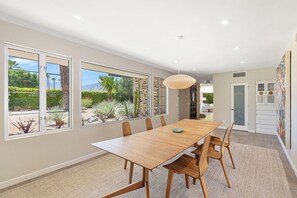 A dining area with a wooden table, chairs, and a view of a landscaped garden through large windows. - The Beehive - A true midcentury estate (Palm Springs)