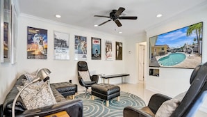 Un salon avec un canapé en cuir noir, un fauteuil noir, une petite table avec une lampe, un ventilateur de plafond et un grand écran de télévision qui diffuse une vue sur la piscine.