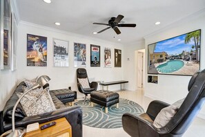 Ein Wohnzimmer mit einem schwarzen Ledersofa, einem schwarzen Sessel, einem kleinen Tisch mit Lampe, einem Deckenventilator und einem großen Fernseher, der Blick ins Schwimmbecken zeigt.