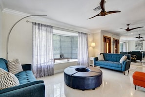 A modern living room with a blue sofa, a round ottoman, and a blue couch. There are ceiling fans, a window with blinds, and a lamp. - Coral Reef Beach House (Lauderdale-by-the-Sea)