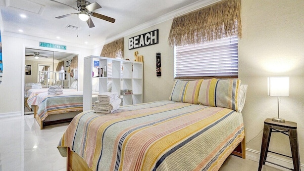 Une chambre avec un lit, un ventilateur de plafond, une fenêtre avec des stores et un panneau mural sur lequel on peut lire le mot « PLAGE ».