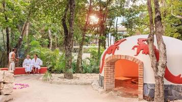 An outdoor area with a brick oven structure, surrounded by trees and greenery.