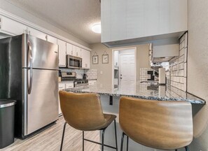 A kitchen with a stainless steel refrigerator, a microwave, and a stove. There are two bar stools and a granite countertop. - Newly Remodeled 3 BR House w/ Fire Pit, 5 min. From Stadium! (Norman)