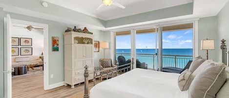 A bedroom with a large bed, a ceiling fan, a view of the ocean, a balcony with chairs, and a dining area with a table and chairs.
