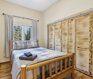 A bedroom with a wooden bed, a window with curtains, a wardrobe with a wooden facade, and a view of trees outside. - The Story Cabin - Processing Equipment & Freezer Storage Coming in 2027 (Soldotna)