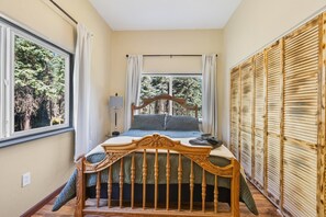 Ein Schlafzimmer mit einem hölzernen Bett, einem Fenster mit Vorhängen, einer Nachttischlampe und Blick auf Bäume.