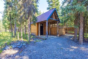 A wooden cabin with a porch, surrounded by trees and gravel ground.