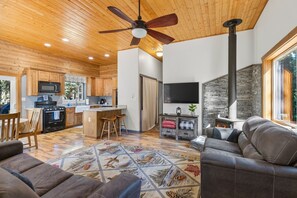 Eine moderne Küche mit Holzschränken, einer zentralen Kochinsel mit Barhocker, einem Essbereich mit Tisch und Stühlen, einem Wohnzimmer mit Sofa und einem Kamin mit Steinwand.