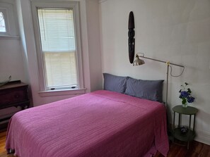A bedroom with a bed, a window with blinds, a bedside lamp, a dresser, and a small table with a vase.