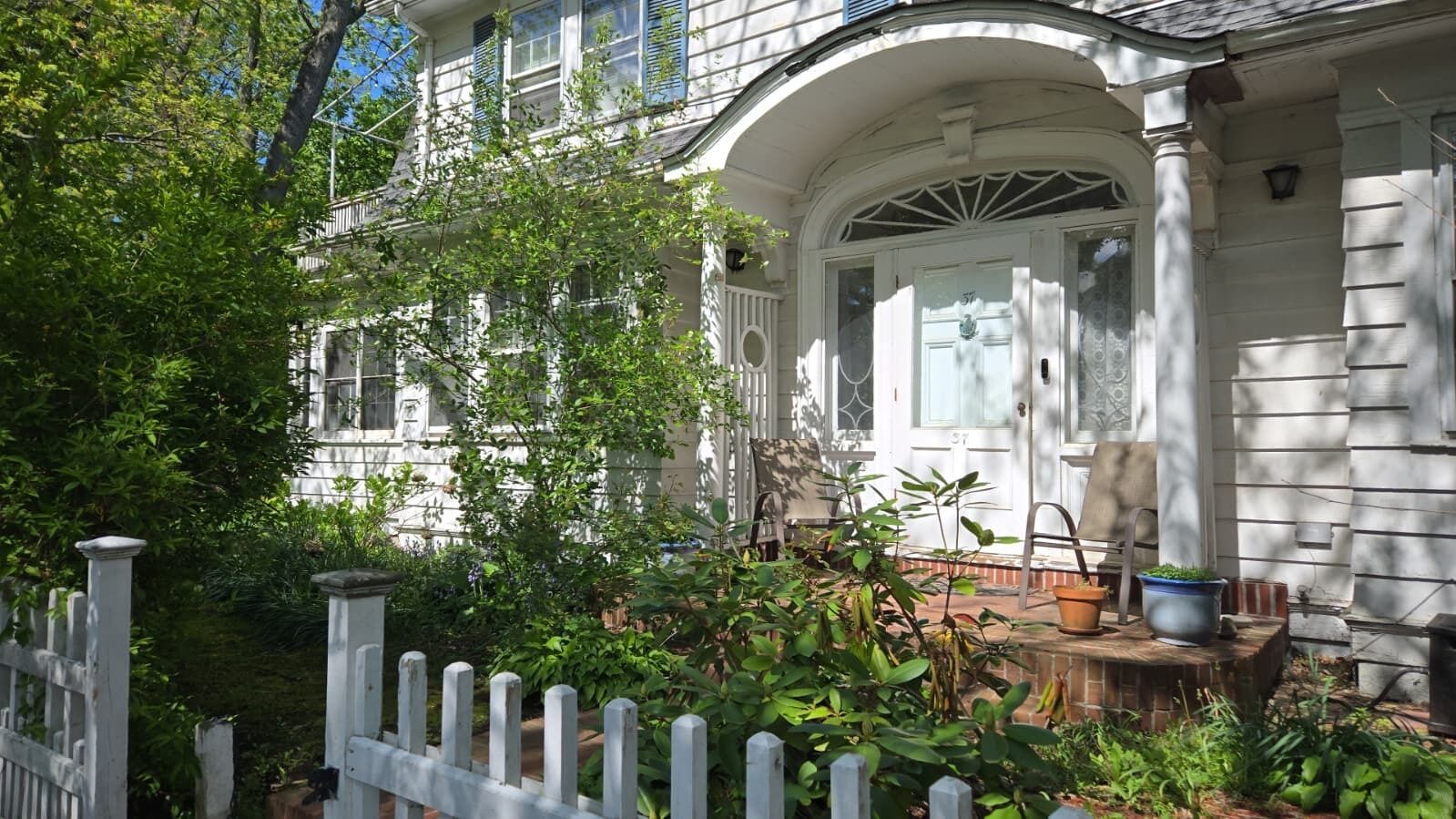 A white house with a porch, a white picket fence, and potted plants.
