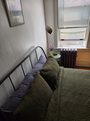 Ein Bett mit Metallgestell, ein Kissen, eine grüne, strukturierte Decke, ein kleiner Tisch mit Blumen in einer Vase und ein Heizkörper.