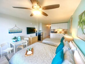 Una habitación de hotel compacta con cama, escritorio con silla, kitchenette y ventilador de techo.