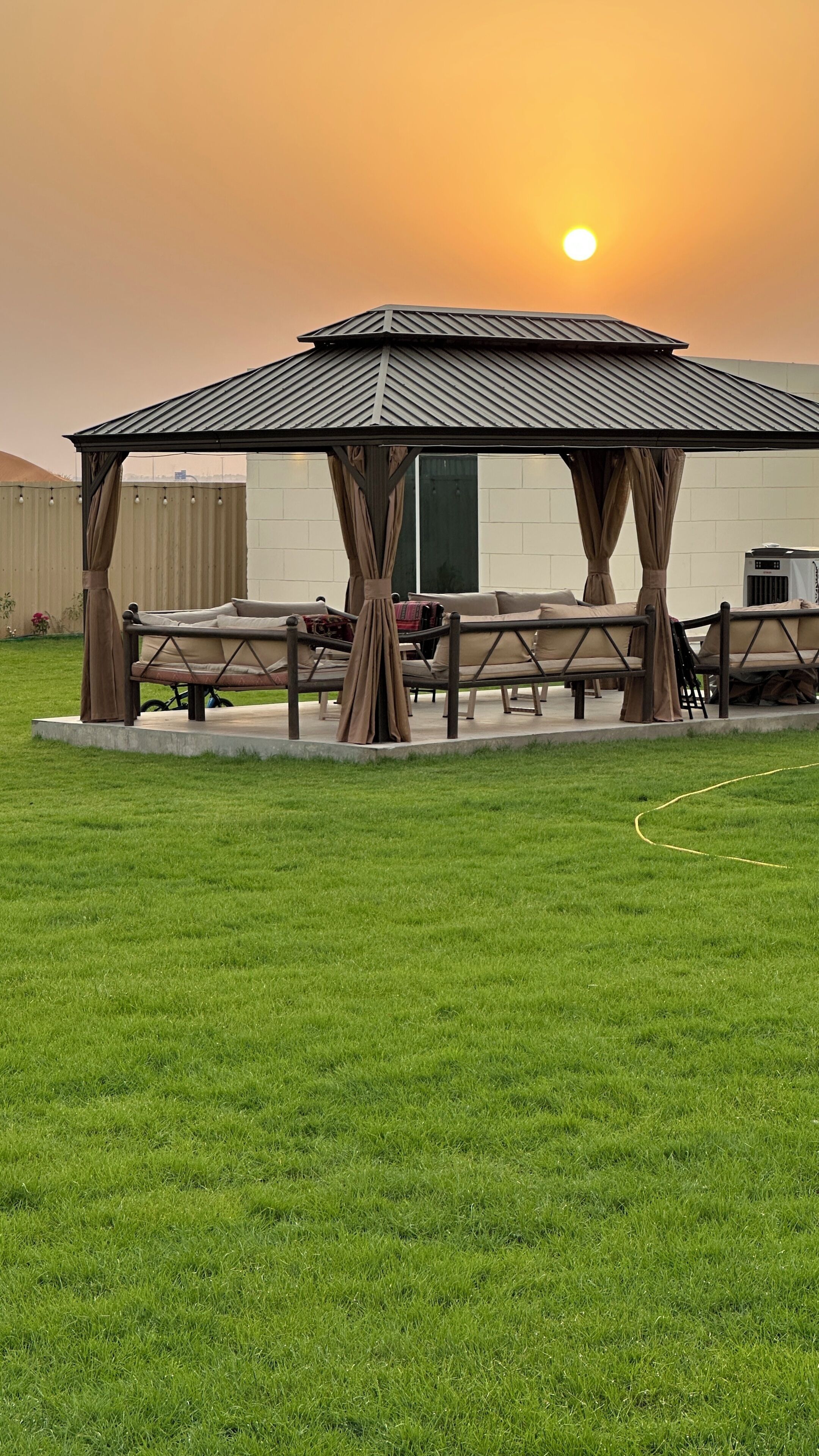 A gazebo with a metal roof and wooden pillars, surrounded by a grassy lawn and a picnic table with benches.