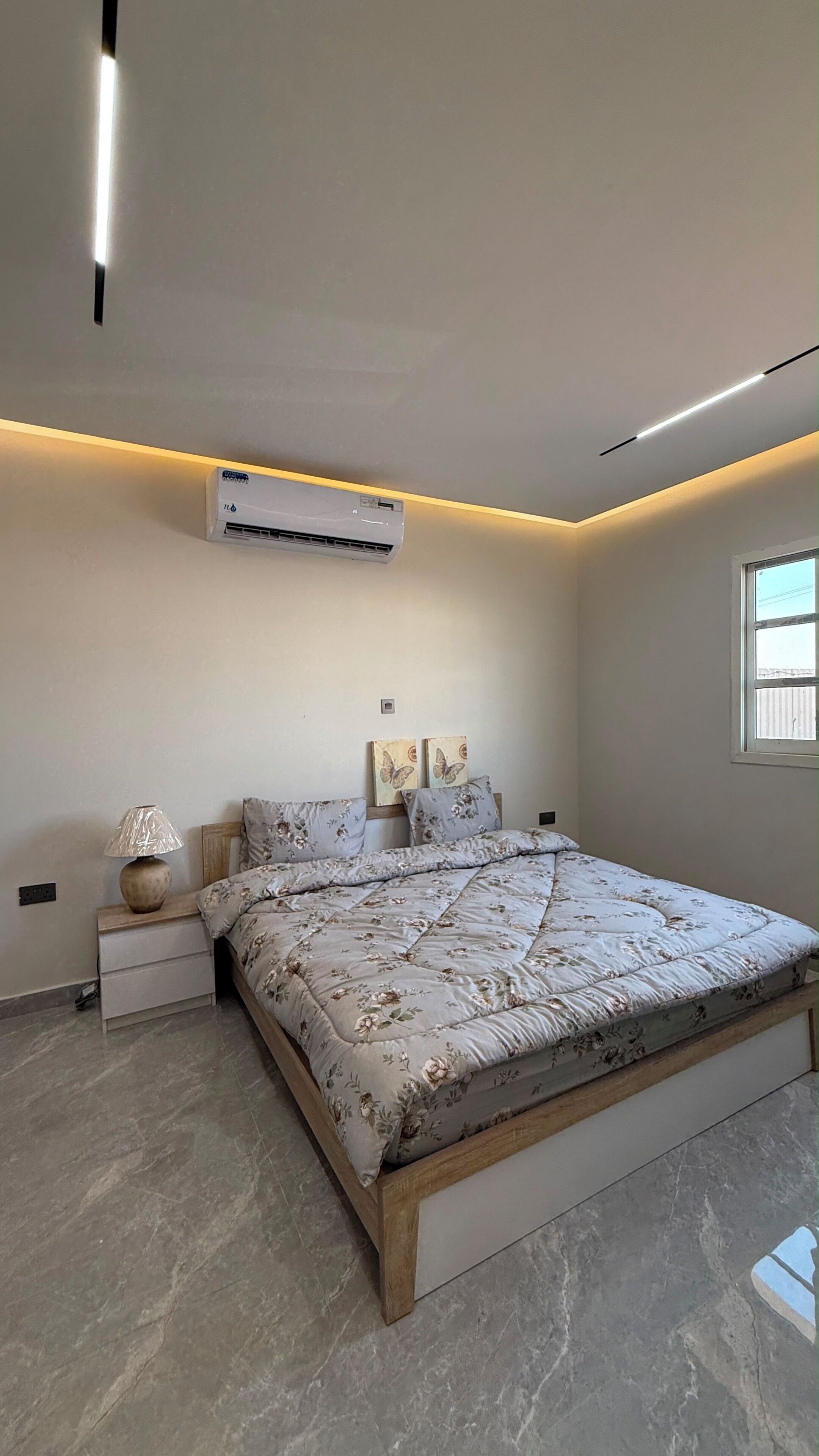 A bedroom with a bed, bedside table, lamp, air conditioning unit, and a window.