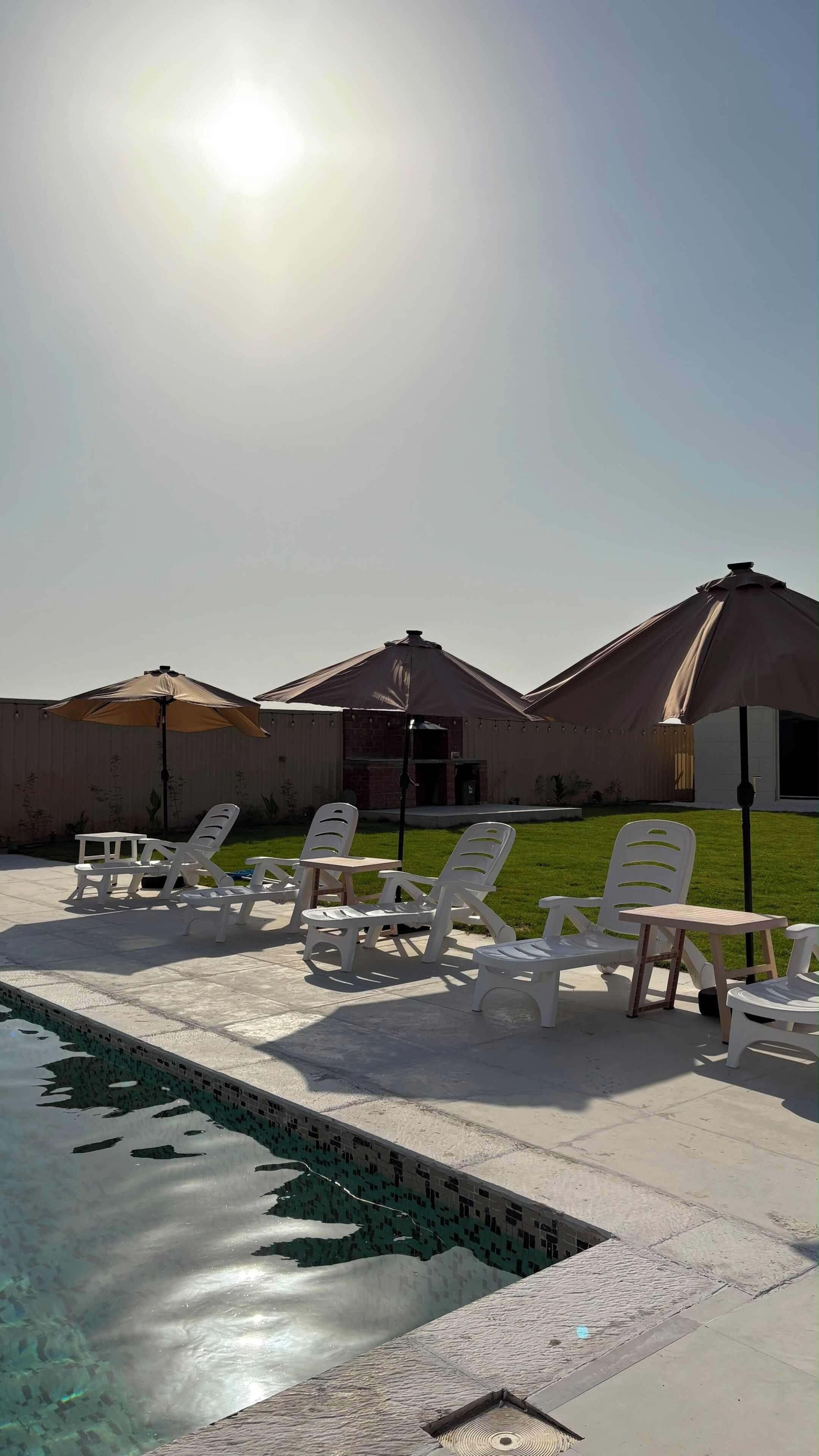 A pool area with lounge chairs and umbrellas, a brick building in the background, and a clear sky.