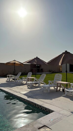 Un área de piscina con sillones de descanso y sombrillas, un edificio de ladrillo al fondo y un cielo despejado.