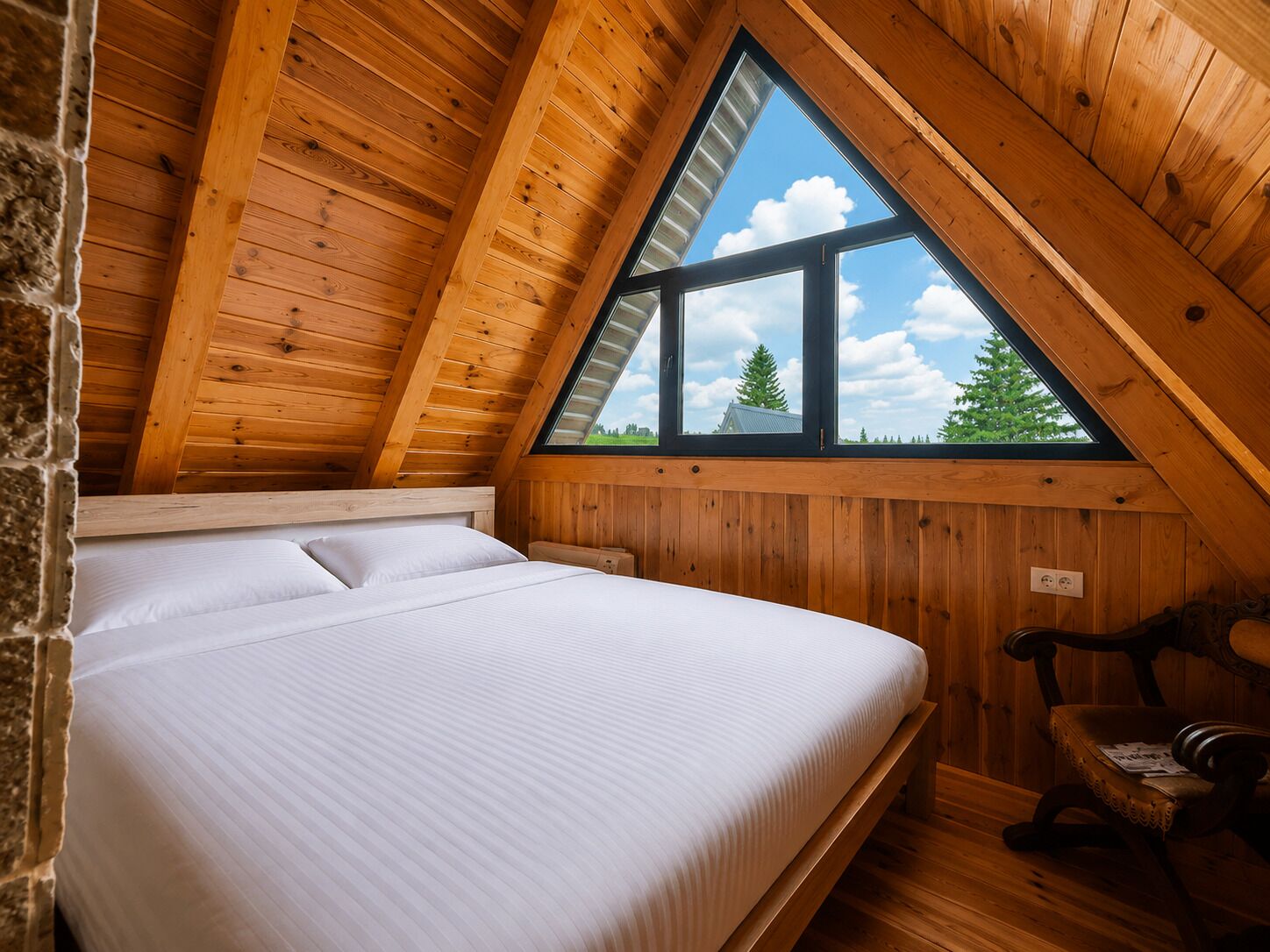 Ein Schlafzimmer mit schrägen Wänden, einem Bett mit weißer Bettwäsche, einem Fenster mit Blick auf Bäume und einem Holzstuhl.