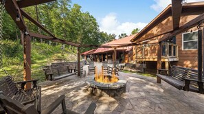 A wooden outdoor patio with stone seating, a fire pit, and a covered area with wooden beams and hanging lights. - Its Got It All Yall by Avantstay Sleeps 27 Indoor Pool + Hot Tub + Theater + Near Pigeon Forge (Pigeon Forge)