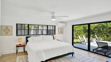 Ein Schlafzimmer mit Bett, Nachttischlampen, Deckenventilator, Fenster mit Jalousien und Blick auf einen Pool und Gartenmöbel.