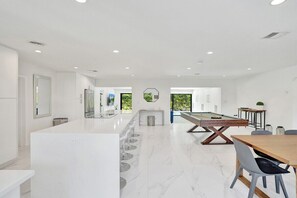 Una cocina moderna con encimera de mármol, mesa de billar y zona de comedor con mesa y sillas de madera.