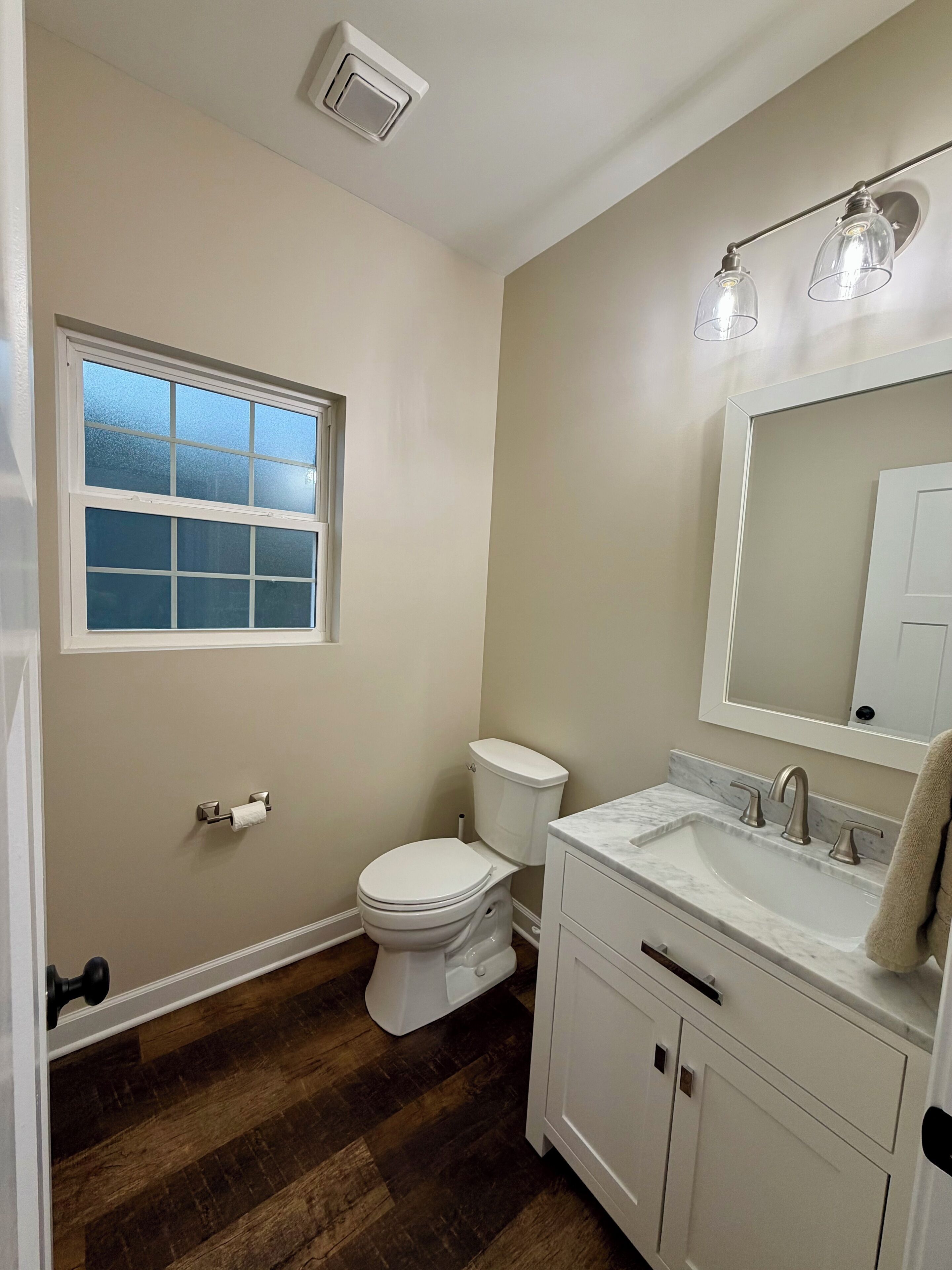 A bathroom with a toilet, sink, mirror, and window.