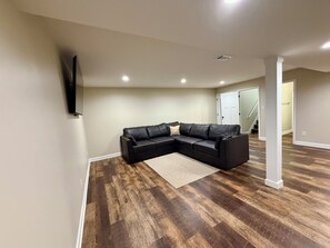 A living room with a sectional sofa, a flat-screen TV mounted on the wall, and a wooden floor.