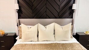 Un lit recouvert d’une couette beige à motifs, avec une tête de lit à chevrons de couleur foncée et une table de chevet surmontée d’une lampe.