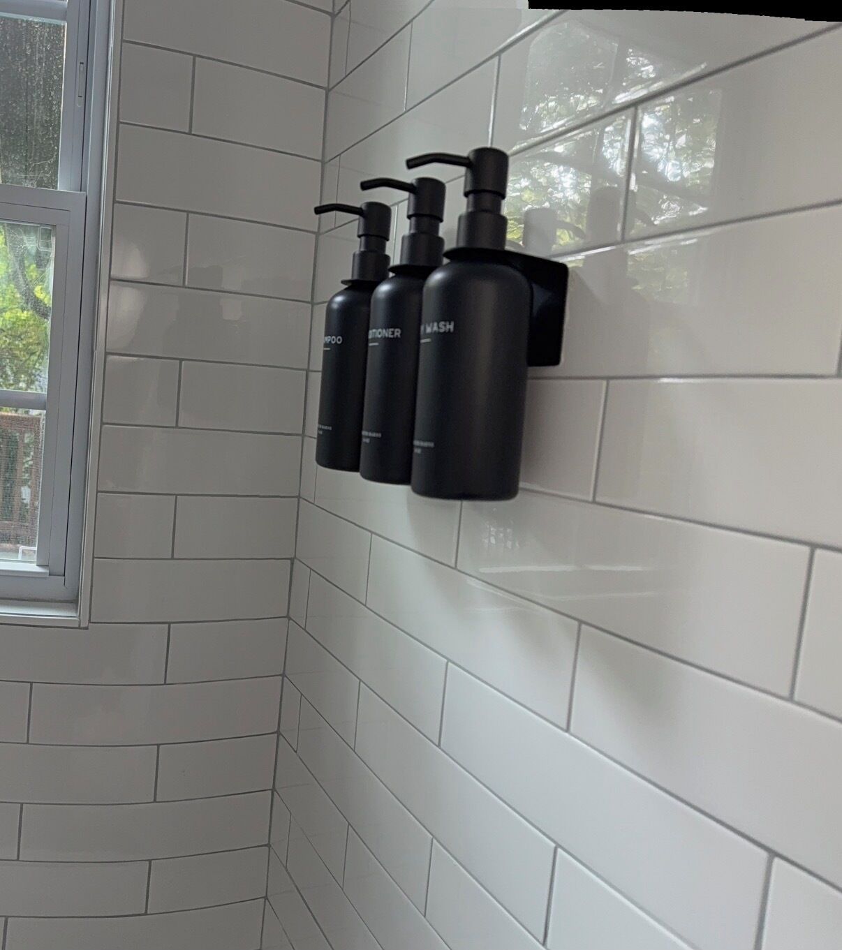 Een badkamer met witte betegelde wanden, een raam en drie zwarte flessen aan de muur.