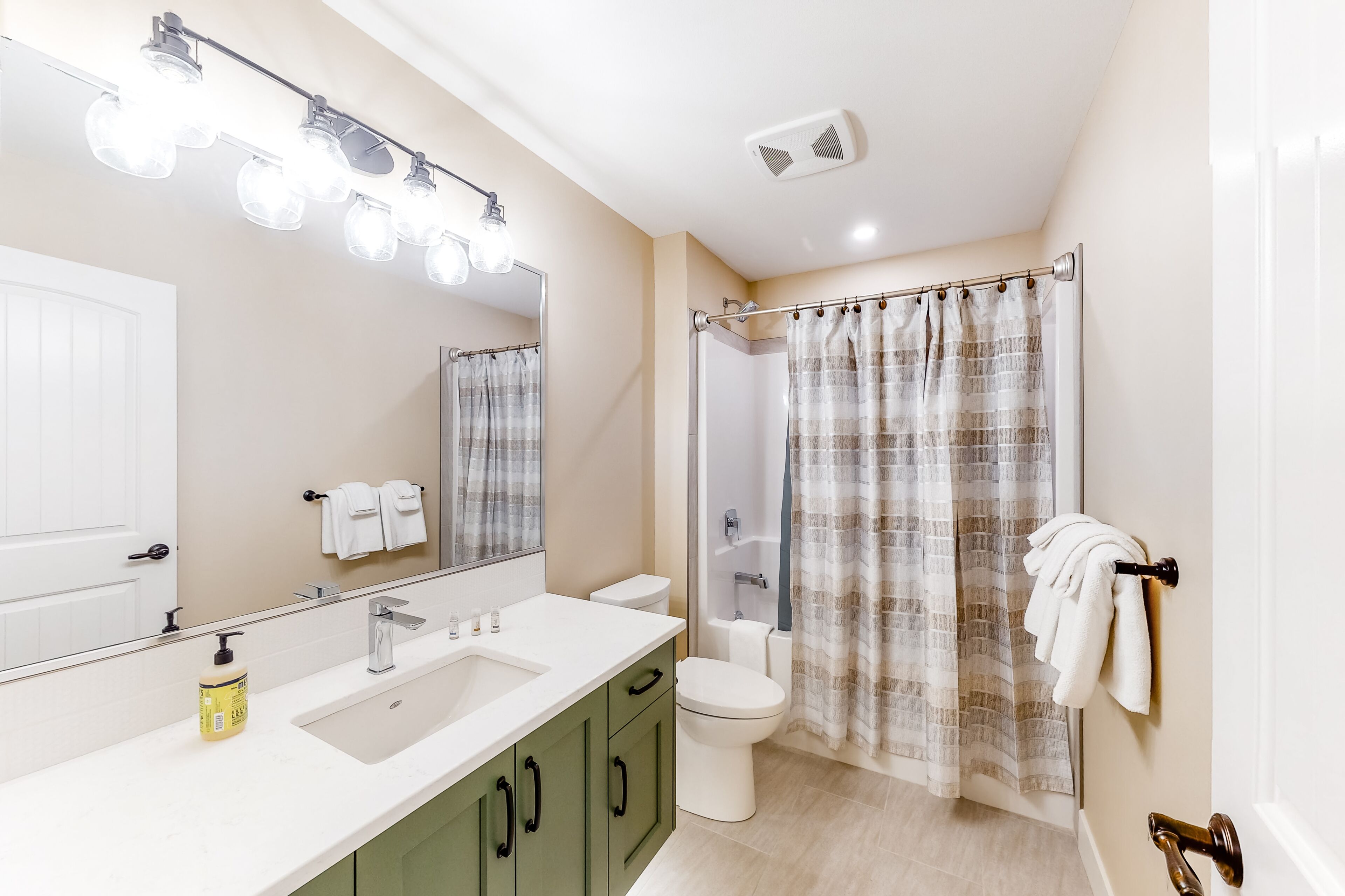 Une salle de bain avec un lavabo blanc, des meubles verts, des toilettes blanches, un rideau de douche et un porte-serviettes.