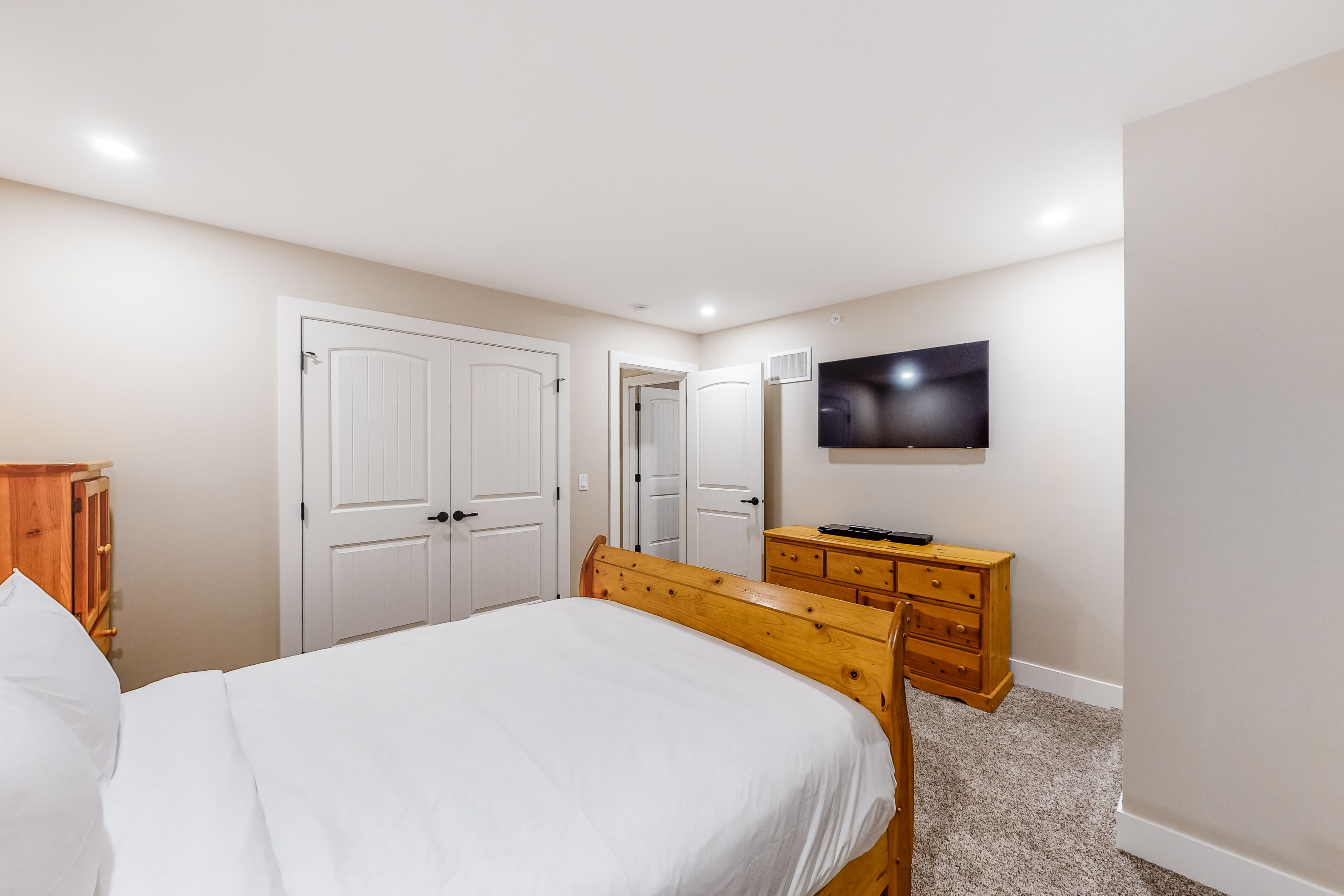 A bedroom with a bed, wooden dresser, wardrobe, and a TV on the wall.