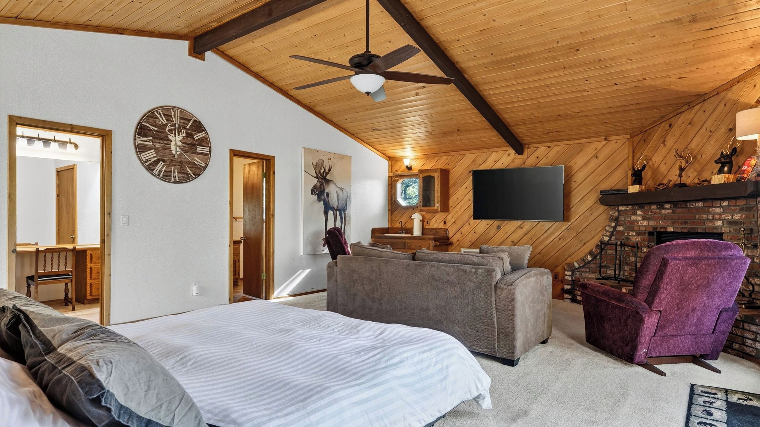 Une chambre à coucher avec un lit, un canapé gris, un fauteuil violet, un plafond en bois, une horloge et une cheminée.