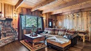 A rustic wooden interior with a fireplace, leather sofa, and a large window overlooking greenery. - Pinewood Perch by Avantstay Layered Decks + Grill + Multiple Living Spaces Slopes + Lake Nearby (Big Bear City)