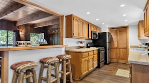 A kitchen with wooden cabinets, a stainless steel refrigerator, and a microwave. There are stools at the counter and a window with curtains. - Pinewood Perch by Avantstay Layered Decks + Grill + Multiple Living Spaces Slopes + Lake Nearby (Big Bear City)