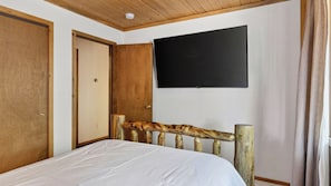 A bedroom with a wooden headboard, a bed, a flat-screen TV mounted on the wall, and a door. - Pinewood Perch by Avantstay Layered Decks + Grill + Multiple Living Spaces Slopes + Lake Nearby (Big Bear City)