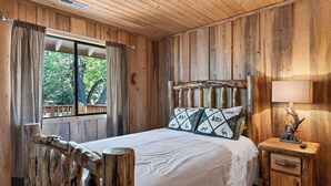Ein Schlafzimmer einer Holzhütte mit Bett, Nachttisch, Lampe und einem Fenster mit Vorhängen.