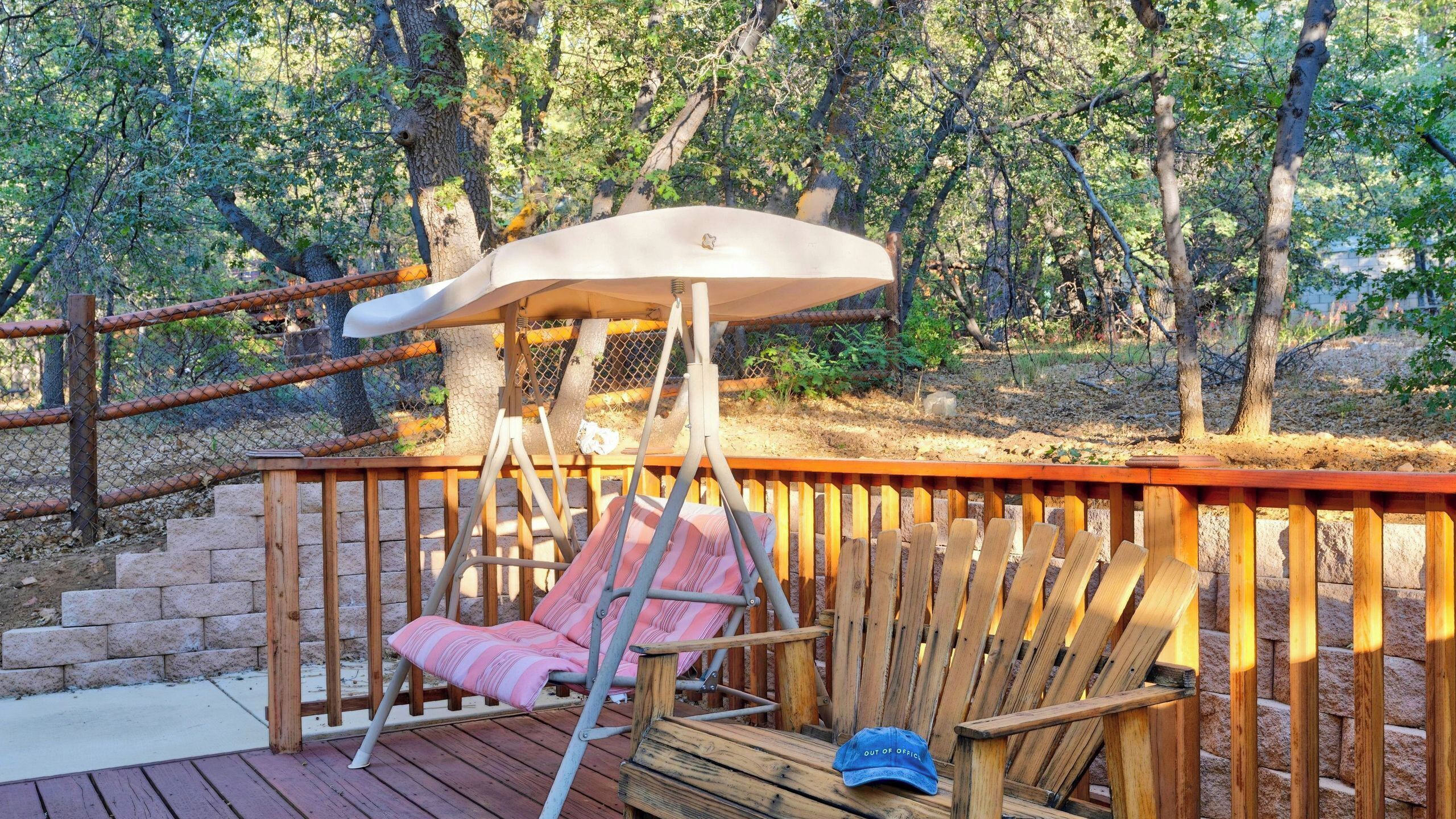 Une terrasse en bois avec une chaise suspendue et un banc, entourée d’une clôture en treillis métallique et d’arbres.