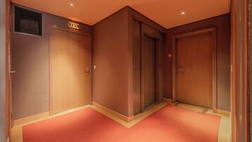 A hotel corridor with two elevators, a red carpet, and a wall-mounted emergency exit sign.