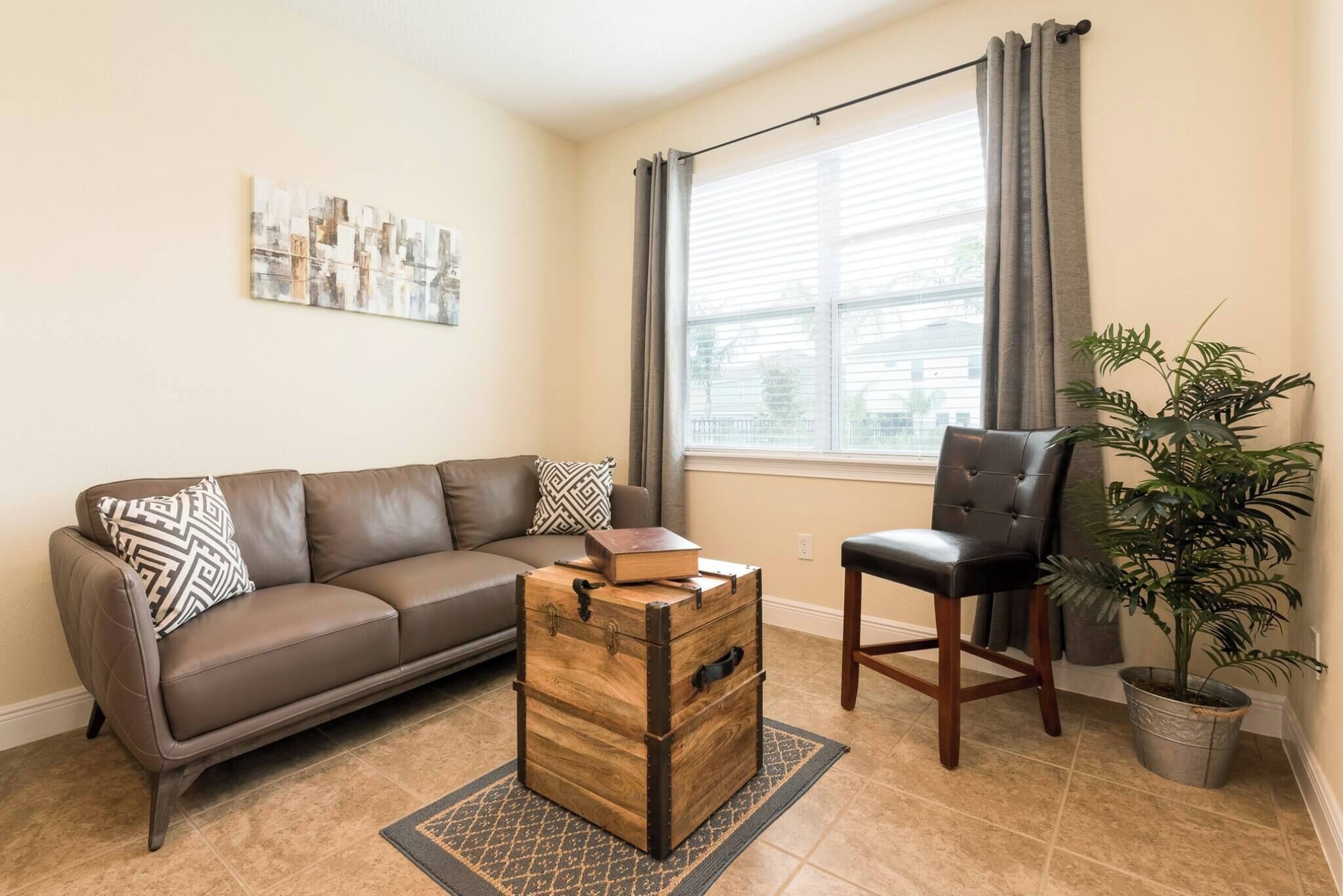 A living room with a brown leather sofa, a wooden chest, a black leather chair, a potted plant, and a window with curtains.