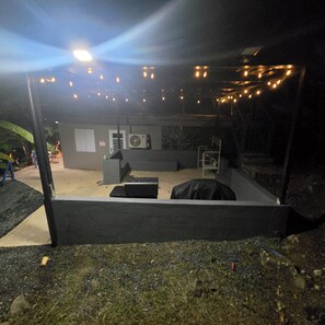 Ein Sitzbereich im Freien mit Tischen und Stühlen, einer kleinen Bühne und Lichterketten.