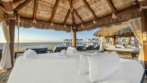 Quarto de hotel à beira-mar com telhado de palha, roupa de cama branca e vista para a praia.