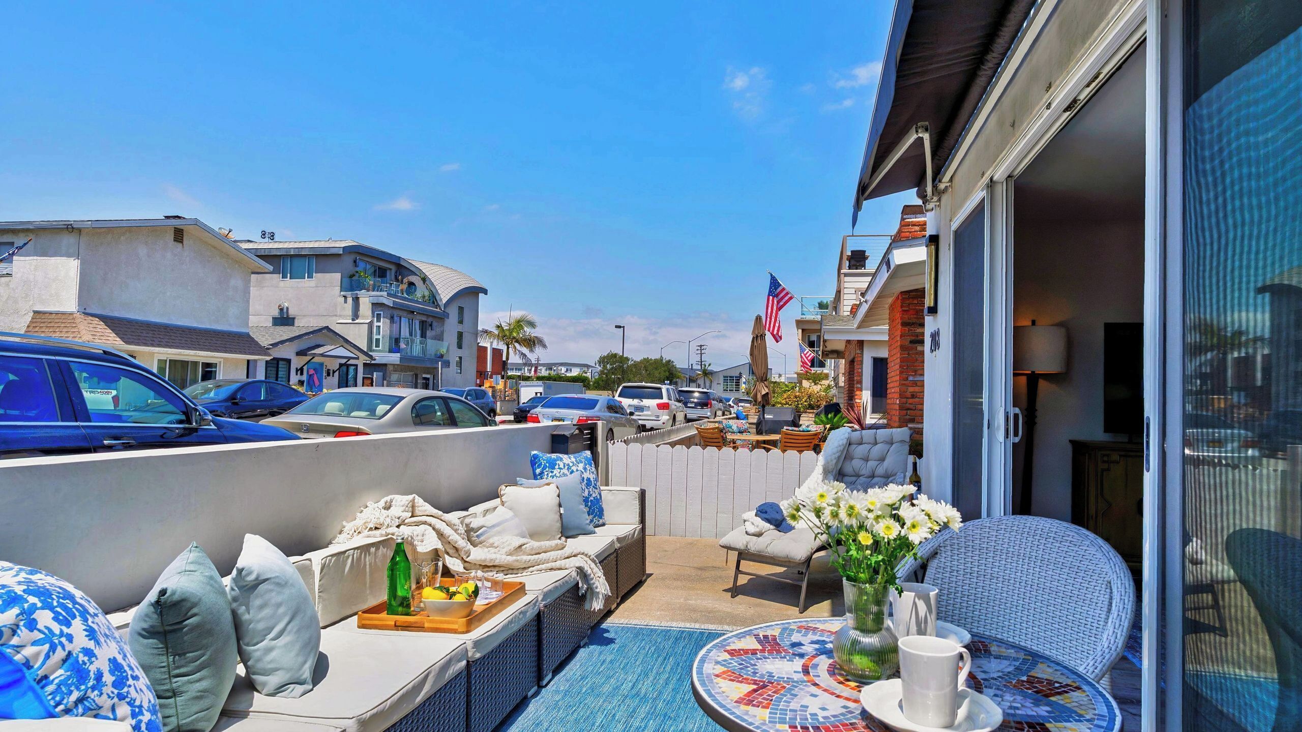 A balcony with outdoor seating, a table with a drink, and a view of a street with parked cars and buildings.