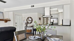 Una cocina moderna con armarios blancos, un refrigerador de acero inoxidable y un comedor con una silla de cuero negro y una mesa dispuesta con platos y un florero con hojas verdes.