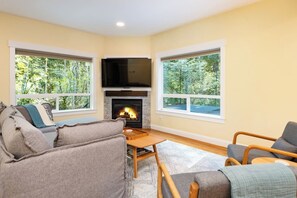A cozy living room with a fireplace, a television, and large windows offering a view of trees. - Forest Retreat | Hot Tub & Sauna (Bellingham)