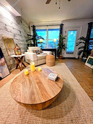 Una sala de estar con una mesa redonda de madera, un sofá, una pequeña mesita auxiliar y una ventana con cortinas.