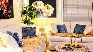 Uma sala de estar com dois sofás brancos, uma mesa de centro redonda, um abajur e um quadro grande com motivos florais na parede.