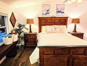 Ein Schlafzimmer mit einem hölzernen Bett, zwei Nachttischlampen, einem Nachttisch, einer Kommode, einem Fenster mit Jalousien und Wandbildern.